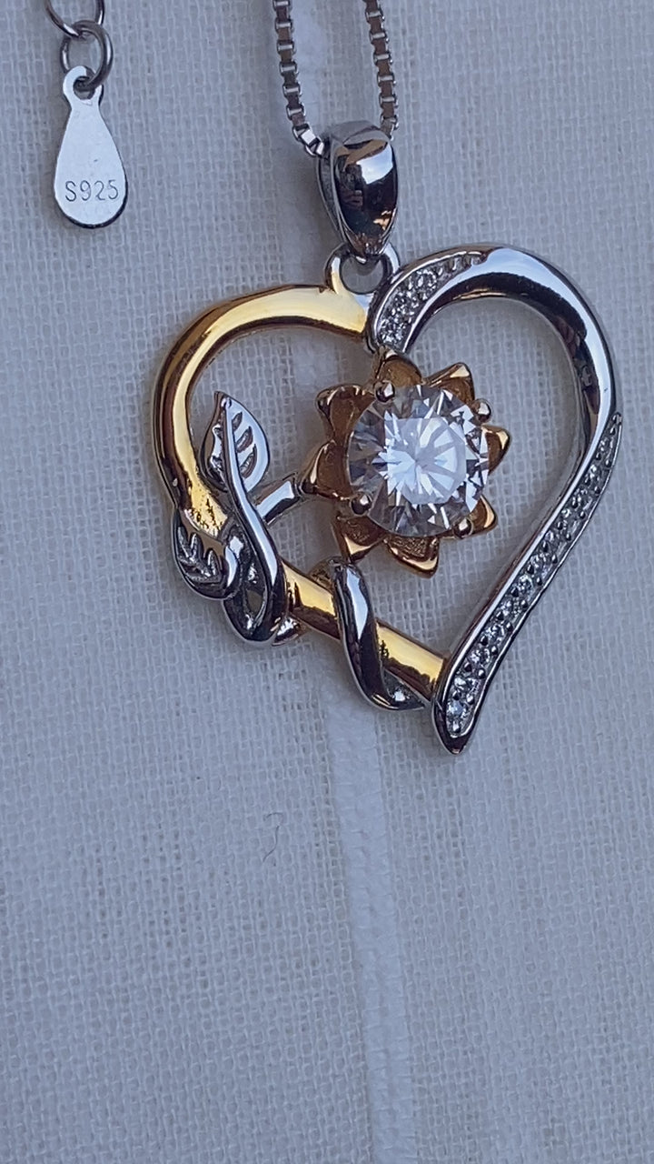 Sterling Silver Heart Pendant with a 1CT Moissanite Diamond, centered in the middle of a golden flower, surrounded by 15 smaller ones, mounted in a prong setting that dangles on a C-link sterling silver chain. 